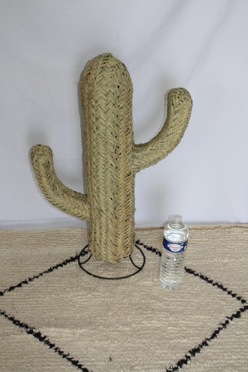 Puede incluir: Una gran escultura de cactus beige tejida con un soporte de metal negro. El cactus tiene dos brazos y est&aacute; de pie sobre una alfombra blanca con ribete negro.