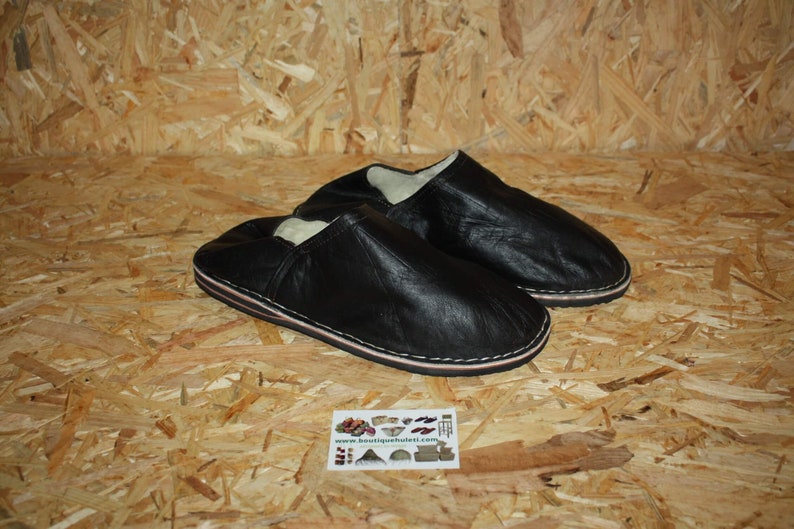 Op de afbeelding: Een paar zwarte leren slippers met een witte voering. De slippers hebben een eenvoudig ontwerp en zijn gemaakt van zacht leer.