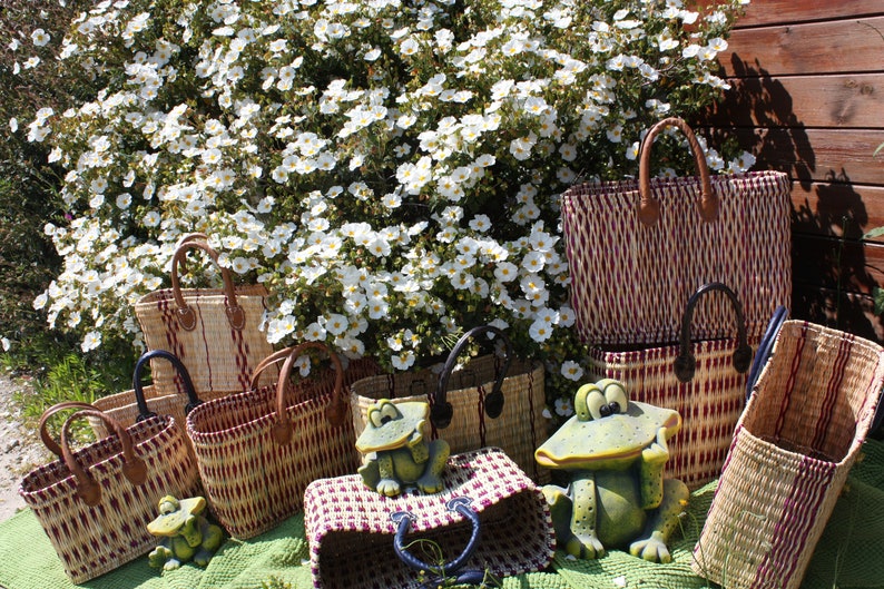 May include: A collection of woven baskets with handles, in various sizes and patterns, with a red and white striped design. The baskets are arranged in front of a bush with white flowers and a wooden wall. Two green frog figurines are placed in front of the baskets.