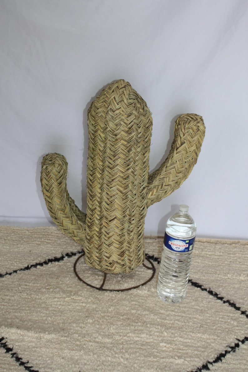 Puede incluir: Una gran escultura de cactus de paja tejida con un soporte de metal. El cactus es de color marr&oacute;n claro y tiene una superficie texturizada. El cactus est&aacute; sobre una alfombra blanca con patrones de diamantes negros.