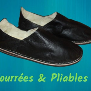 Op de afbeelding: Een paar zwarte leren slippers met een witte bontvoering. De slippers zijn gevouwen en hebben een gestikte naad rond de rand. De tekst "Fourr&eacute;es & Pliables!" staat in het groen onder de slippers.