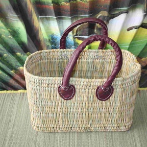 May include: A woven basket with two burgundy leather handles. The basket is made of natural fibres and has a rectangular shape.