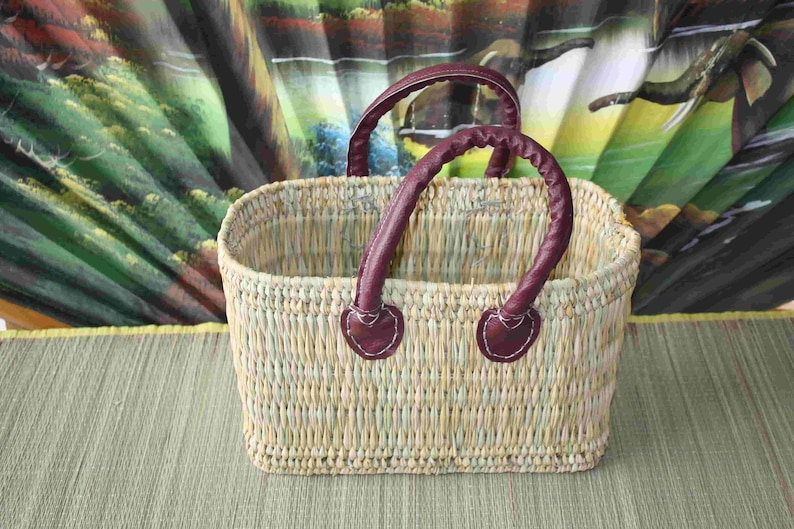 May include: A woven basket with two burgundy leather handles. The basket is made of natural fibres and has a rectangular shape.