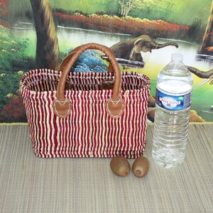 May include: A red and white striped woven basket with brown leather handles. The basket is sitting on a brown surface with two kiwi fruits and a clear plastic water bottle.