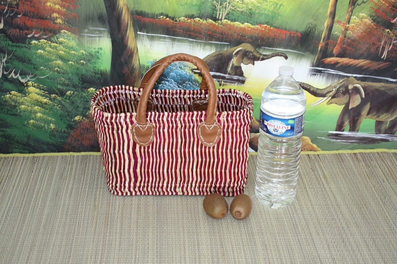 May include: A red and white striped woven basket with brown leather handles. The basket is sitting on a brown surface with two kiwi fruits and a clear plastic water bottle.