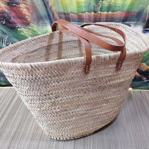 May include: A large, natural woven basket with brown leather handles. The basket is made of a light beige straw and has a wide, oval shape.