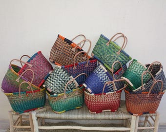 May include: A collection of woven tote bags in various colors, including green, blue, red, and purple. Each bag has a rectangular shape with a woven pattern and a pair of brown handles. The bags are stacked on a wooden bench against a white wall.
