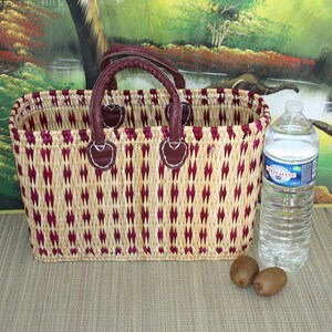 May include: A woven basket with a burgundy and natural colour pattern. The basket has two leather handles and is sitting on a woven mat. There are two kiwi fruits and a clear plastic water bottle in the image.