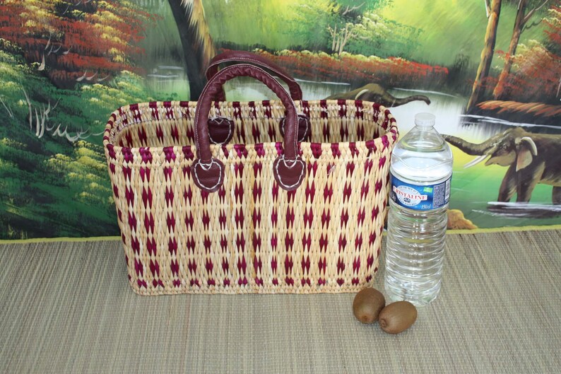 May include: A woven basket with a burgundy and natural colour pattern. The basket has two leather handles and is sitting on a woven mat. There are two kiwi fruits and a clear plastic water bottle in the image.