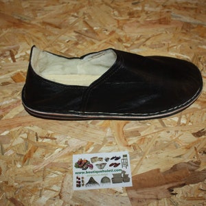 Op de afbeelding: Een zwarte leren pantoffel met een witte voering. De pantoffel ligt op een houten oppervlak.