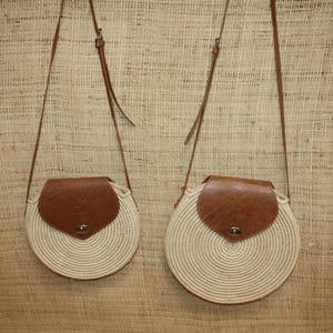 May include: Two round straw bags with brown leather flaps and straps. The bags have a woven design and a circular shape.