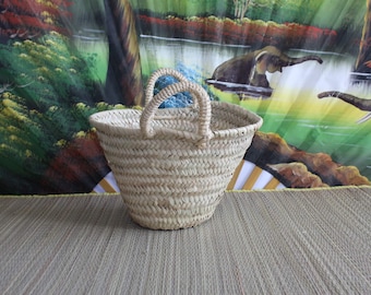 Op de afbeelding: Een geweven strooien mand met twee handvatten. De mand is lichtbruin en heeft een afgeronde vorm. Hij staat op een geweven mat.