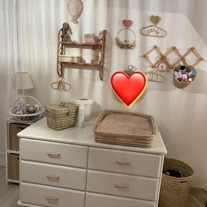 Op de afbeelding: Een witte commode met zes laden en gouden handvatten. De commode is versierd met een verschoningsmat, een rieten mand en een lamp. De muur achter de commode is versierd met een luchtballon, een hart en een regenboog.