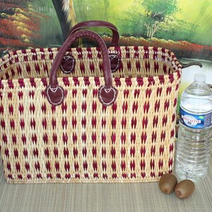 May include: A woven basket with red and beige stripes and leather handles. The basket is sitting on a beige surface with two kiwi fruits and a clear plastic water bottle.