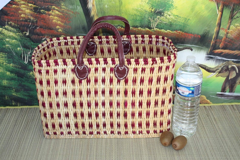 May include: A woven basket with red and beige stripes and leather handles. The basket is sitting on a beige surface with two kiwi fruits and a clear plastic water bottle.