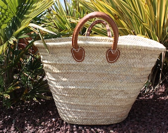 May include: A large, woven basket with brown leather handles. The basket is made of natural straw and has a light beige color. The basket is sitting on a bed of small, red rocks.