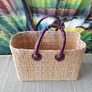 May include: A rectangular woven basket with brown leather handles. The basket is made of natural materials and has a light brown colour.