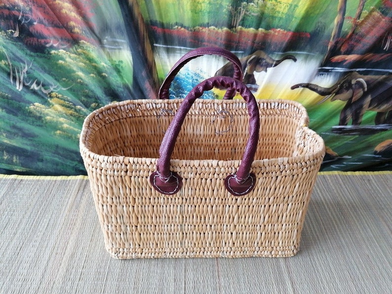 May include: A rectangular woven basket with brown leather handles. The basket is made of natural materials and has a light brown colour.