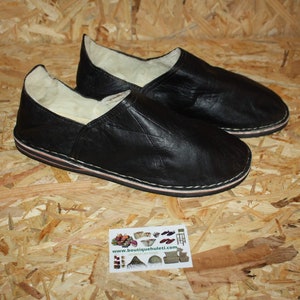Op de afbeelding: Een paar zwarte leren slippers met een witte schapenvacht voering. De slippers hebben een eenvoudig ontwerp en een platte zool.