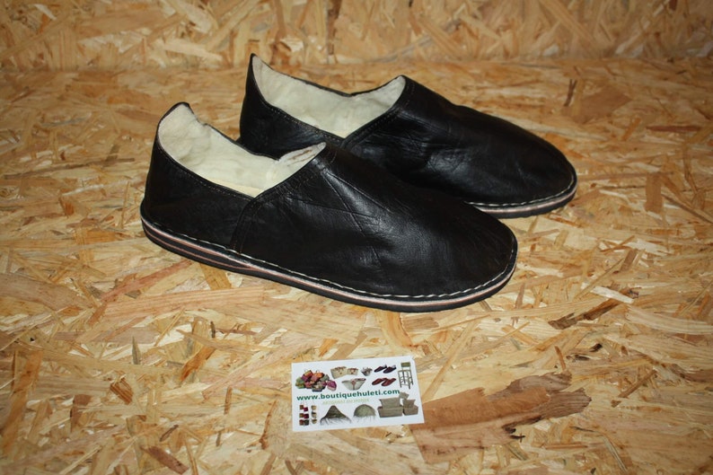 Op de afbeelding: Een paar zwarte leren slippers met een witte schapenvacht voering. De slippers hebben een eenvoudig ontwerp en een platte zool.