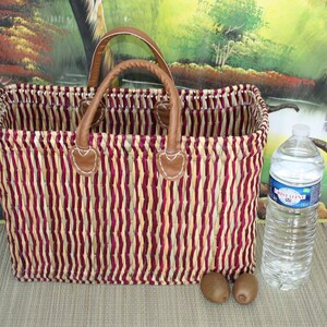 May include: A striped woven basket with brown leather handles and a red and cream colour scheme. The basket is sitting on a brown surface with a bottle of water and two kiwi fruits.