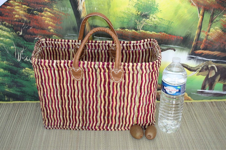 May include: A striped woven basket with brown leather handles and a red and cream colour scheme. The basket is sitting on a brown surface with a bottle of water and two kiwi fruits.