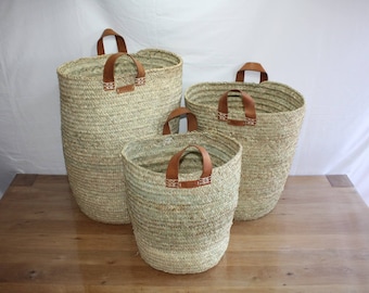 May include: Three woven seagrass baskets with leather handles. The baskets are different sizes and are arranged in a row on a wooden surface.