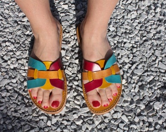 May include: A pair of colourful leather sandals with a multi-coloured design. The sandals have a turquoise, yellow, and red leather strap design. The sandals are on a gravel surface.