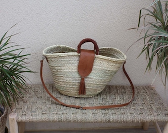 Pode incluir: Uma grande bolsa de palha tecida com uma alça de couro marrom e uma longa alça de couro marrom. A bolsa está sentada em um banco tecido.