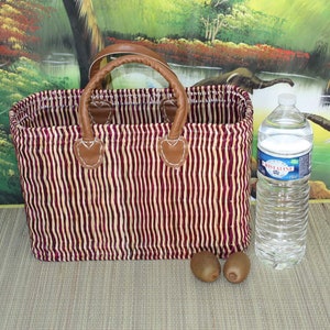 May include: A red and white striped woven basket with brown leather handles. The basket is sitting on a brown surface with two kiwi fruits and a clear plastic water bottle.