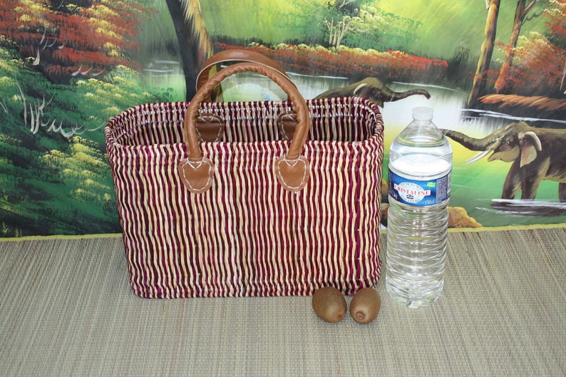 May include: A red and white striped woven basket with brown leather handles. The basket is sitting on a brown surface with two kiwi fruits and a clear plastic water bottle.