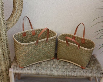 May include: Two woven straw baskets with leather handles. The baskets are different sizes and are sitting on a woven bench.