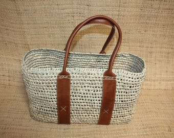Puede incluir: Un bolso tote tejido con asas y detalles de cuero marrón. El bolso está hecho de un material claro, de tejido abierto, con forma rectangular. Las asas están sujetas con correas de cuero marrón, sobre un fondo texturizado.