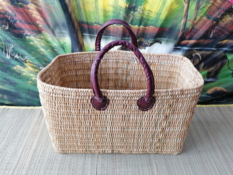 May include: A rectangular woven basket with two brown leather handles. The basket is made of natural materials and has a light brown colour.