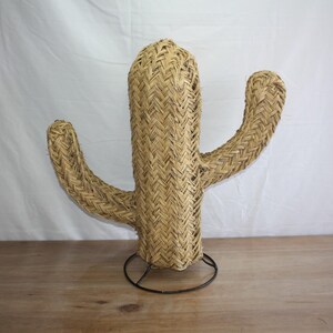 Puede incluir: Una gran escultura de cactus tejida de color beige con una base de metal negro. El cactus tiene un tallo central alto y dos brazos m&aacute;s peque&ntilde;os.