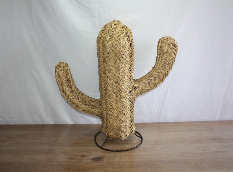 Puede incluir: Una gran escultura de cactus tejida de color beige con una base de metal negro. El cactus tiene un tallo central alto y dos brazos m&aacute;s peque&ntilde;os.