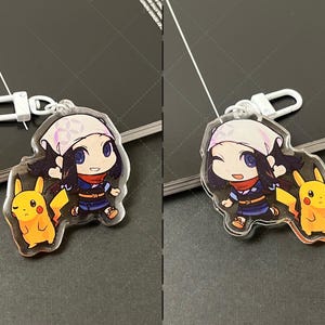 May include: A clear acrylic keychain featuring a cartoon character with dark hair, a light-colored head covering, and a red scarf, standing next to a yellow Pikachu character. The keychain includes a white plastic lobster clasp attachment.