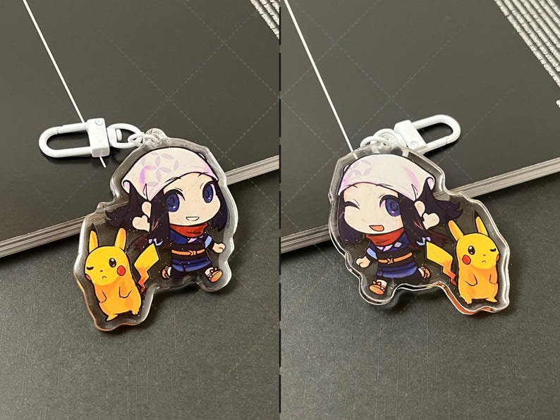 May include: A clear acrylic keychain featuring a cartoon character with dark hair, a light-colored head covering, and a red scarf, standing next to a yellow Pikachu character. The keychain includes a white plastic lobster clasp attachment.