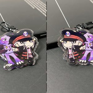 May include: A double-sided acrylic keychain featuring a cartoon character in a dark uniform with a peaked cap and a purple creature with sharp claws. The keychain has a black metal lobster clasp and a glittery finish over the printed artwork.