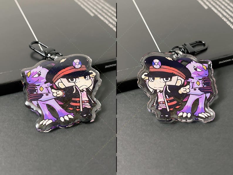 May include: A double-sided acrylic keychain featuring a cartoon character in a dark uniform with a peaked cap and a purple creature with sharp claws. The keychain has a black metal lobster clasp and a glittery finish over the printed artwork.