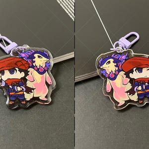 May include: A clear acrylic keychain featuring a chibi-style character wearing a red cap and scarf, standing next to a bipedal creature with purple flames around its neck. The charm includes a light purple lobster clasp for attachment.