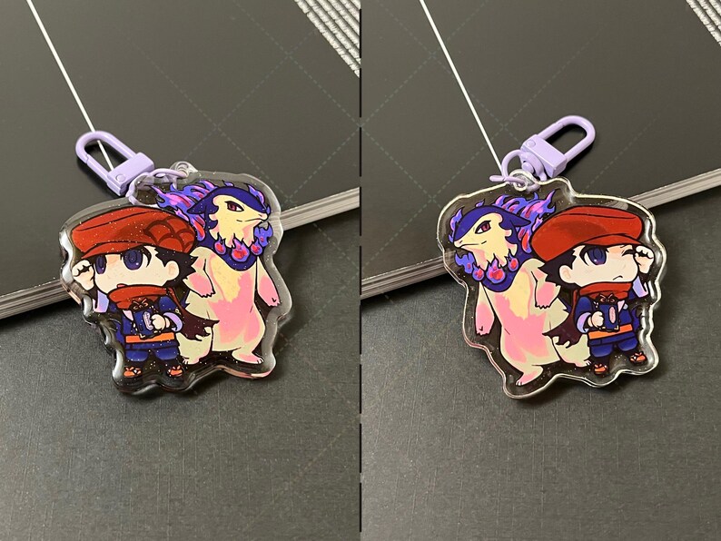 May include: A clear acrylic keychain featuring a chibi-style character wearing a red cap and scarf, standing next to a bipedal creature with purple flames around its neck. The charm includes a light purple lobster clasp for attachment.