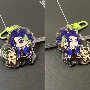 May include: A clear acrylic keychain featuring a cartoon character with dark blue hair and a small yellow creature with green accents. The character wears a blue and black outfit with a hexagonal pendant. A bright lime green plastic clasp is attached.