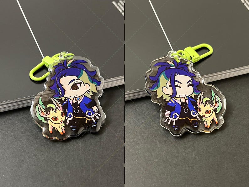 May include: A clear acrylic keychain featuring a cartoon character with dark blue hair and a small yellow creature with green accents. The character wears a blue and black outfit with a hexagonal pendant. A bright lime green plastic clasp is attached.