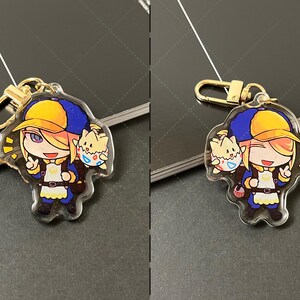 May include: An acrylic keychain featuring a cartoon character with blonde hair wearing a yellow and blue cap, a blue jacket, and a white apron, accompanied by a small creature in a patterned eggshell. The charm includes a gold-toned metal clasp.
