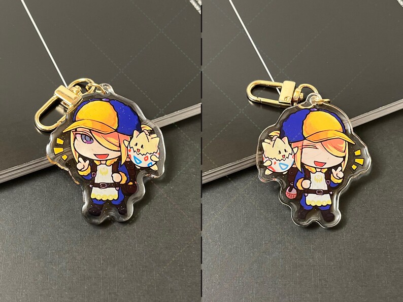 May include: An acrylic keychain featuring a cartoon character with blonde hair wearing a yellow and blue cap, a blue jacket, and a white apron, accompanied by a small creature in a patterned eggshell. The charm includes a gold-toned metal clasp.