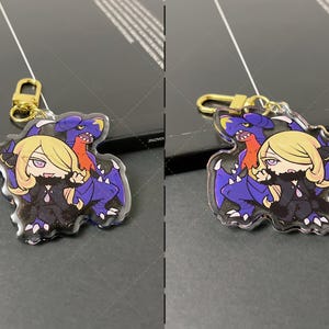 May include: A clear acrylic keychain featuring a chibi-style illustration of a blonde character with purple eyes and a dark outfit standing in front of a large, purple and blue dragon-like creature. The charm includes a gold-colored metal lobster clasp.