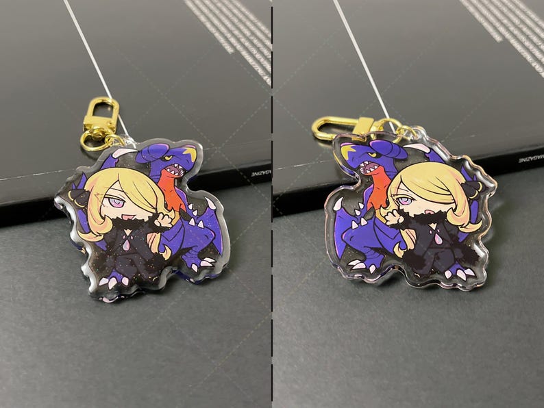 May include: A clear acrylic keychain featuring a chibi-style illustration of a blonde character with purple eyes and a dark outfit standing in front of a large, purple and blue dragon-like creature. The charm includes a gold-colored metal lobster clasp.