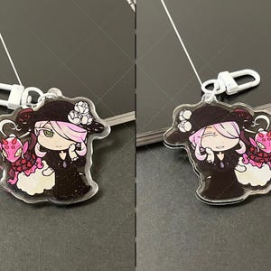 May include: A clear acrylic keychain featuring a character with light pink hair wearing a black hat with white flowers and a black dress, accompanied by a small pink dragon-like creature with red spots, attached to a white plastic lobster clasp.