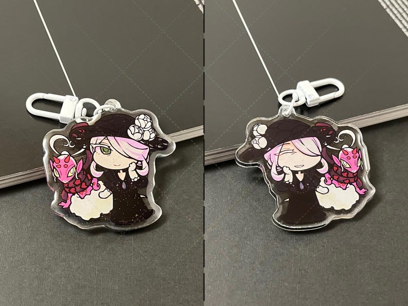 May include: A clear acrylic keychain featuring a character with light pink hair wearing a black hat with white flowers and a black dress, accompanied by a small pink dragon-like creature with red spots, attached to a white plastic lobster clasp.